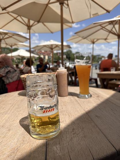 Ein Glas mit „Würstlkuchel“-Bier steht auf einem Holztisch im Freien. Im Hintergrund sind weitere Gläser, eine Senf-Flasche und ein unscharfes Bild von Personen unter großen Sonnenschirmen zu sehen. Die Szene spielt in einem Biergarten oder einer Außengastronomie bei sonnigem Wetter. Der Himmel ist blau mit einigen Wolken.