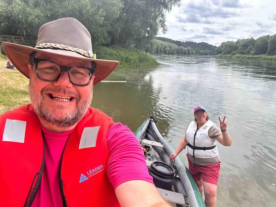 Zwei Personen in einem grünen Schlauchlanadier auf einem ruhigen Fluss. Die Person im Vordergrund trägt eine rote Schwimmweste mit der Aufschrift „LEADER ACCESSORIES“, eine Brille und einen beigen Hut mit geflochtenem Band. Die Person im Hintergrund trägt eine weiße Schwimmweste, eine rote Shorts und eine Basecap mit pinkem Muster. Sie zeigt das Peace-Zeichen mit den Fingern. Im Hintergrund sind grüne Bäume und ein bewölkter Himmel zu sehen. Die Szene wirkt entspannt und abenteuerlich.