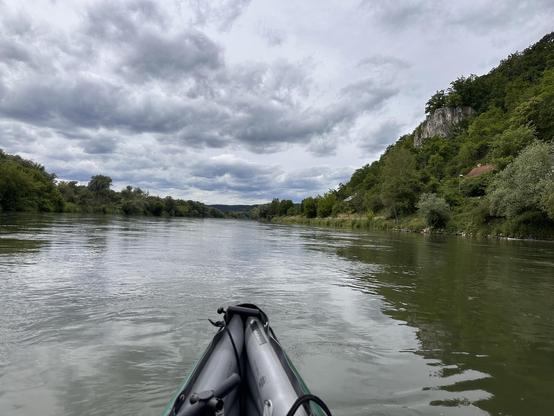 Eine Perspektive aus einem Kajak auf einem breiten Fluss. Im Vordergrund sind die Spitze des Kajaks und ein Teil des Paddels zu sehen. Der Fluss ist ruhig und von grünen, bewaldeten Ufern gesäumt. Am rechten Ufer ragt ein steiler, felsiger Hügel mit Bäumen und Sträuchern empor. Der Himmel ist mit dunklen Wolken bedeckt, was auf ein herannahendes Gewitter oder Regen hindeutet. Die Stimmung wirkt abenteuerlich und naturnah.