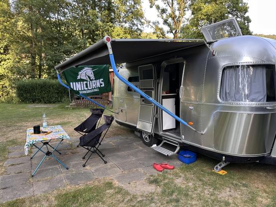 Ein silberner Airstream-Wohnwagen steht auf einem Campingplatz mit gepflastertem Boden. An der Seite des Wohnwagens ist ein blauer Sonnenschutz befestigt, der Schatten spendet. Vor dem Wohnwagen steht ein Klapptisch mit einer bunten Tischdecke, auf dem eine Wasserflasche und ein schwarzer Gegenstand (möglicherweise ein Lautsprecher oder eine Lampe) zu sehen sind. Daneben steht ein klappbarer Campingstuhl. An der Seite des Wohnwagens hängt eine grüne Flagge mit dem Logo der Schwäbisch Hall Unico…
