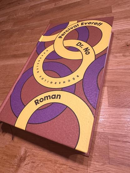 Ein Buch mit dem Titel ‚Percival Everett – Dr. No‘ liegt auf einem Holztisch. Der Umschlag ist in warmen Farben (Rot, Gelb, Violett) gestaltet und zeigt ein abstraktes, kreisförmiges Design. Der Autor Percival Everett und der Titel ‚Dr. No‘ sind in schwarzer Schrift auf dem Umschlag zu lesen. Unten steht das Wort ‚Roman‘ in großer, schwarzer Schrift. Das Logo der Büchergilde Gutenberg ist ebenfalls auf dem Umschlag zu erkennen. Das Buch wirkt hochwertig und künstlerisch gestaltet.