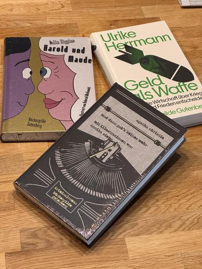 Drei Bücher liegen auf einem Holztisch:
 1. Vorderes Buch: Ein Buch mit dem Titel ‚Agatha Christie und dann gab’s keines mehr‘ mit Illustrationen von Annika Siems. Der Umschlag ist schwarz-weiß und zeigt eine stilisierte Zeichnung einer Gruppe von Figuren, die um einen Tisch sitzen. Das Buch ist als Kriminalroman der Büchergilde Gutenberg gekennzeichnet.
 2. Mittleres Buch: ‚Harold und Maude‘ von Colin Higgins, veröffentlicht von der Büchergilde Gutenberg. Der Umschlag zeigt zwei stilisierte Ge…