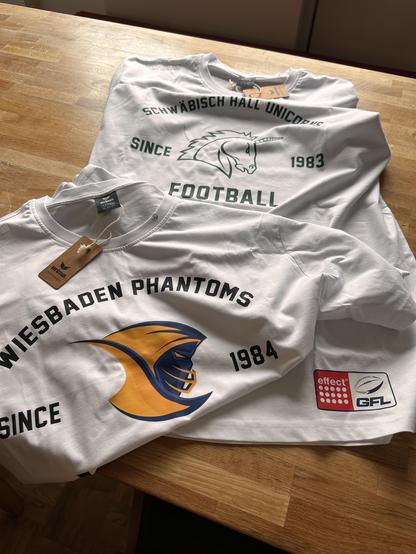 Zwei weiße T-Shirts liegen auf einem Holztisch.
- Oberes T-Shirt:
- Aufdruck: „SCHWÄBISCH HALL UNICORNS“ in grünen Buchstaben.
- darunter: „SINCE 1983“ und ein stilisiertes Einhorn-Logo.
- am unteren Rand: „FOOTBALL“ in grünen Buchstaben.
- Marke: Erima (Etikett am Kragen).
- rechts: Ein Aufnäher mit dem Logo der GFL (German Football League) und dem Schriftzug „effect“.

- Unteres T-Shirt:
- Aufdruck: „WIESBADEN PHANTOMS“ in schwarzen Buchstaben.
- darunter: Ein stilisiertes Logo mit einem oran…