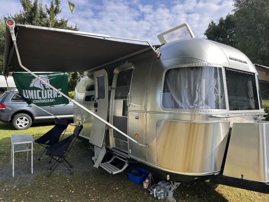 Ein silberner Airstream-Wohnwagen steht auf einem Campingplatz mit gepflastertem Boden. An der Seite des Wohnwagens ist ein schwarzer Sonnenschutz befestigt, der Schatten spendet. Vor dem Wohnwagen steht ein Klapptisch mit zwei klappbaren Campingstühlen. An der Seite des Wohnwagens hängt eine grüne Flagge mit dem Logo der Schwäbisch Hall Unicorns (ein stilisiertes Einhorn). Im Hintergrund sind ein graues Auto, grüne Bäume und ein weiterer Wohnwagen zu erkennen. Die Szene wirkt entspannt und som…