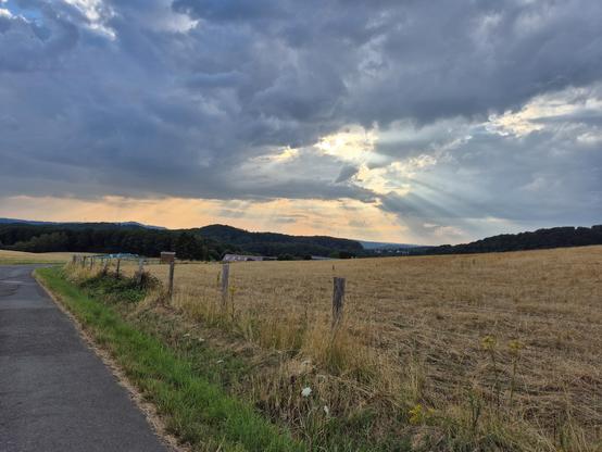 Vor Sonnenuntergang auf einer Höhe des Naturpark Saar-Hunsrück. Sonnenstrahlen brechen magisch durch dunkle Wolkendecke. Links Teil eines schmalen asphaltierten Feldweges, rechts Stoppelfeld (gemähltes Getreidefeld, zum Feldweg durch Stacheldtahtzaun begrenzt ), im Hintergrund bewaldete Berge, davor sind die Dächer eines Bauernhofs zu sehen.