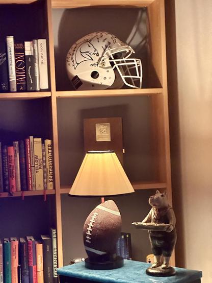 Ein Regal mit verschiedenen Gegenständen und Dekorationen:
- Oberstes Fach: Ein weißer American-Football-Helm der Unicorns mit grünen Aufklebern und Unterschriften.
- Mittleres Fach: Eine Tischlampe mit einem Schirm in Beige und einem Sockel in Form eines braunen American-Footballs.
- Rechts daneben: Eine kleine Figur eines Nilpferds, das ein Tablett hält.
- Linkes Fach: Mehrere Bücher mit verschiedenen Titeln, darunter „Faicon“, „Yellow Face“, „Echtzeitalter“ und weitere.
- Unterstes Fach: Wei…