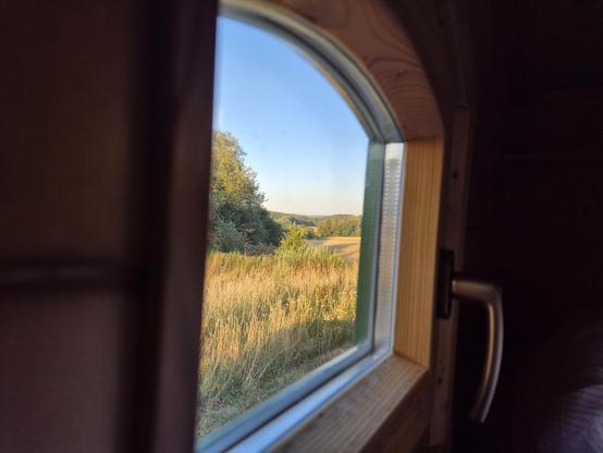 Blick durch ein kleines Holzfenster mit Alugriff aus einem Schäferwagen am Morgen. Panorama mit trockenen, hellgrün-strohgelben Wiesen und Wäldchen (Büsche, vorwiegend Laubbäume) links und am Horizont. Hellblauen, wolkenloser Himmel. Um das Fenster die Wand ist wie das Fenster aus hellem Fichtenholz. Das Bild ist liegend in einem Etagenbett (unten) aufgenommen.
