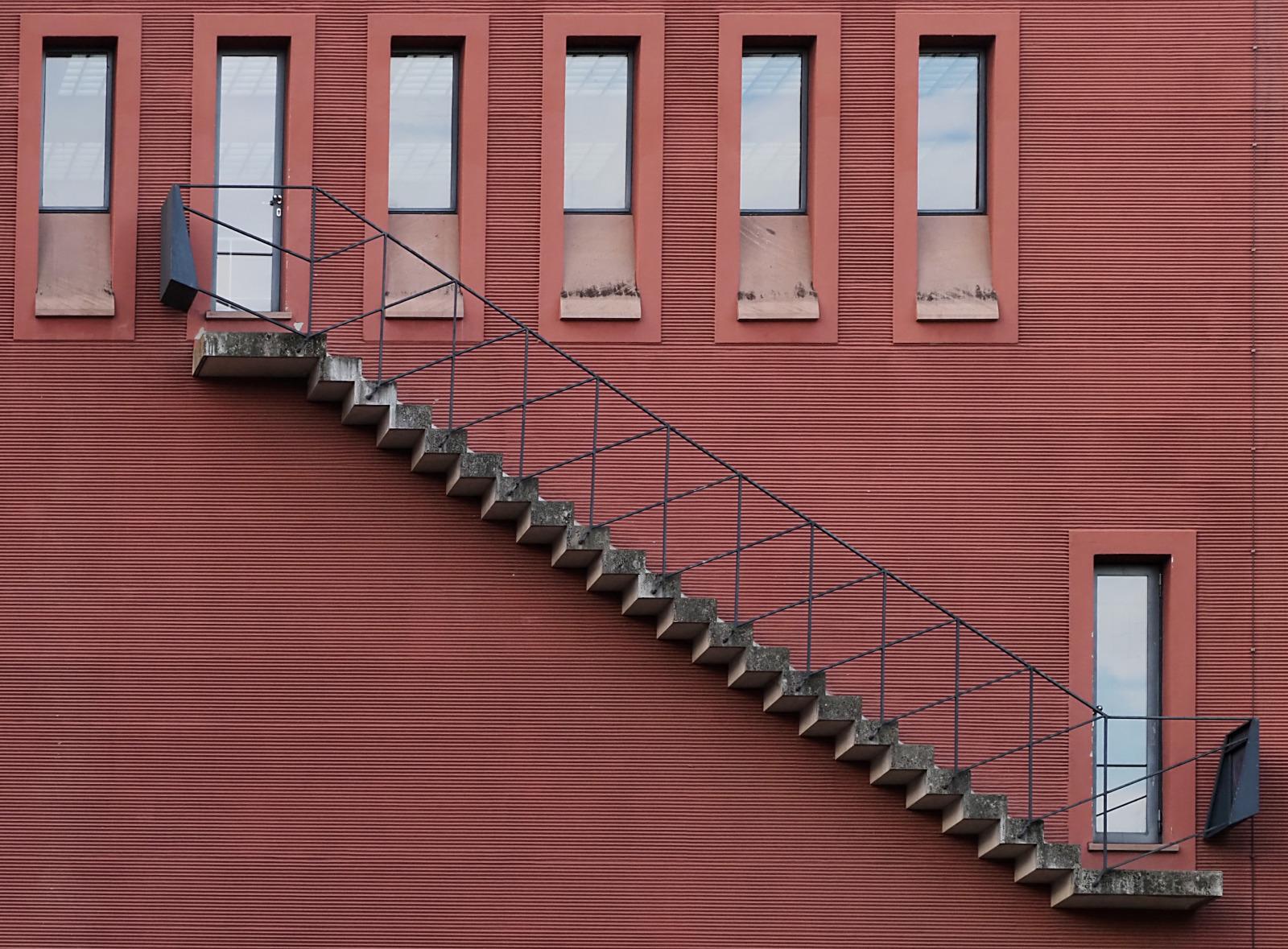 Rot verputzte Wand mit Fensterreihe oben und schmaler Tür rechts unten, verbunden durch eine Außentreppe.