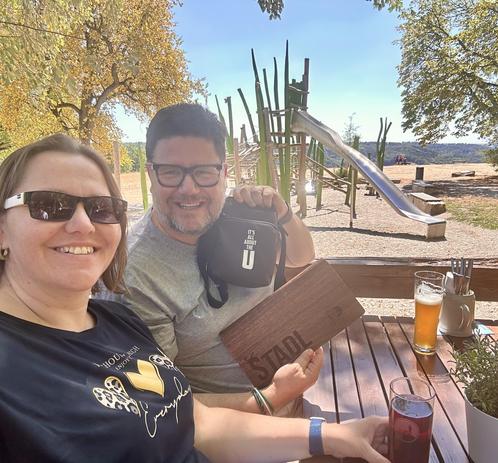 „Zwei Personen sitzen an einem Holztisch im Freien und halten ein holzbeschriftetes Brett mit der Aufschrift ‚STADL‘ in die Kamera.
- Vordere Person: Trägt ein schwarzes T-Shirt mit einem goldenen Logo und einer Sonnenbrille.
- Hintere Person: Trägt ein graues T-Shirt und eine schwarze Tasche mit der Aufschrift ‚It’s all about the U‘.
- Auf dem Tisch: Zwei Gläser mit Getränken – eines mit hellen Bier und eines mit dunklem Getränk (möglicherweise ein weiteres Bier oder ein Softdrink).
- Hintergr…