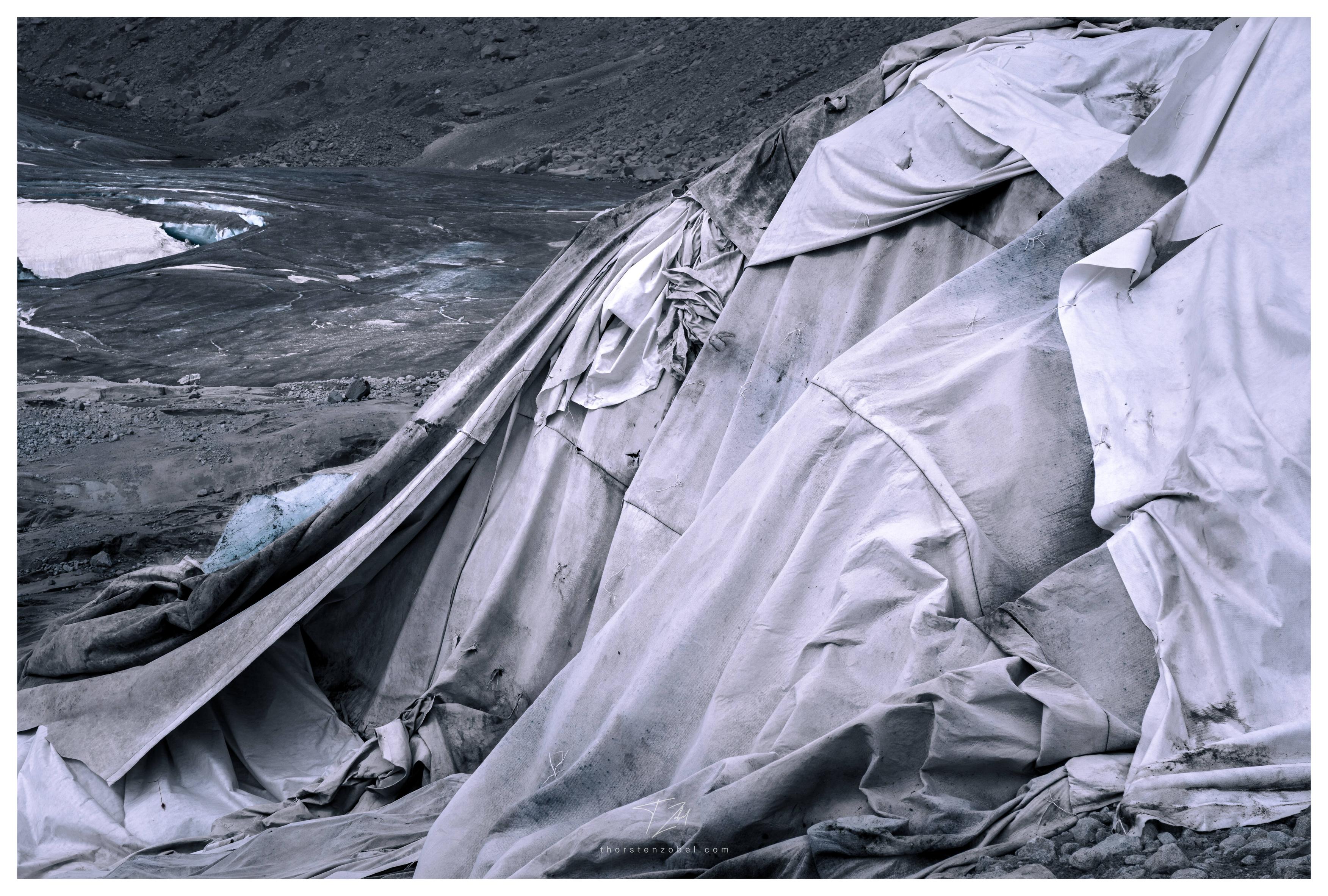 White tarps cover the ice of a glacier. In the background, the terminus of a glacier tongue is visible. The photo has a dystopian atmosphere due to its almost black-and-white, high-contrast processing.