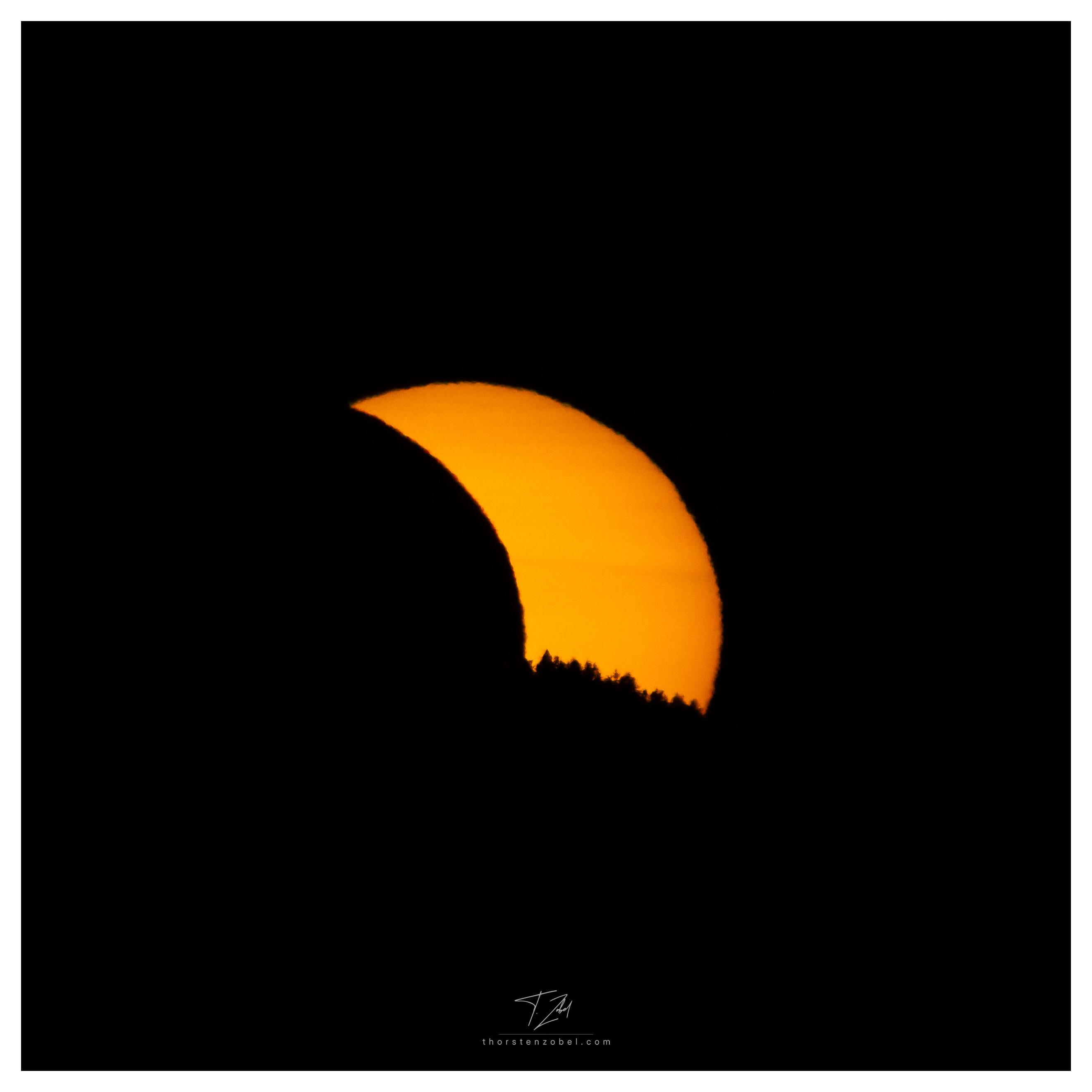The sun sets as a red-orange crescent behind a mountain. Trees are visible as black shadows at the bottom of the sun on the ridge. The crescent is caused by a partial solar eclipse.