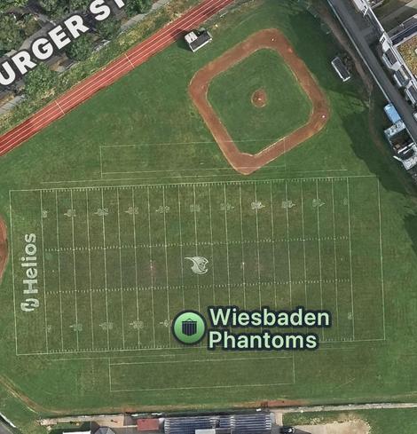 Eine Luftaufnahme eines American-Football-Feldes mit Markierungen und Logos.
- Feld: Das Spielfeld ist grün mit weißen Linien für die Yard-Markierungen (10, 20, 30, 40, 50, etc.). In der Mitte des Feldes ist das Logo der Wiesbaden Phantoms (ein stilisierter Phantom-Kopf) zu sehen.
- Die Endzone trägt den Namen „Helios“ in weißer Schrift.
- Umgebung: Oberhalb des Feldes befindet sich ein Baseball-Feld mit braunem Infield.
Unten rechts steht „Wiesbaden Phantoms“.
Die Szene zeigt die Sportanlage a…