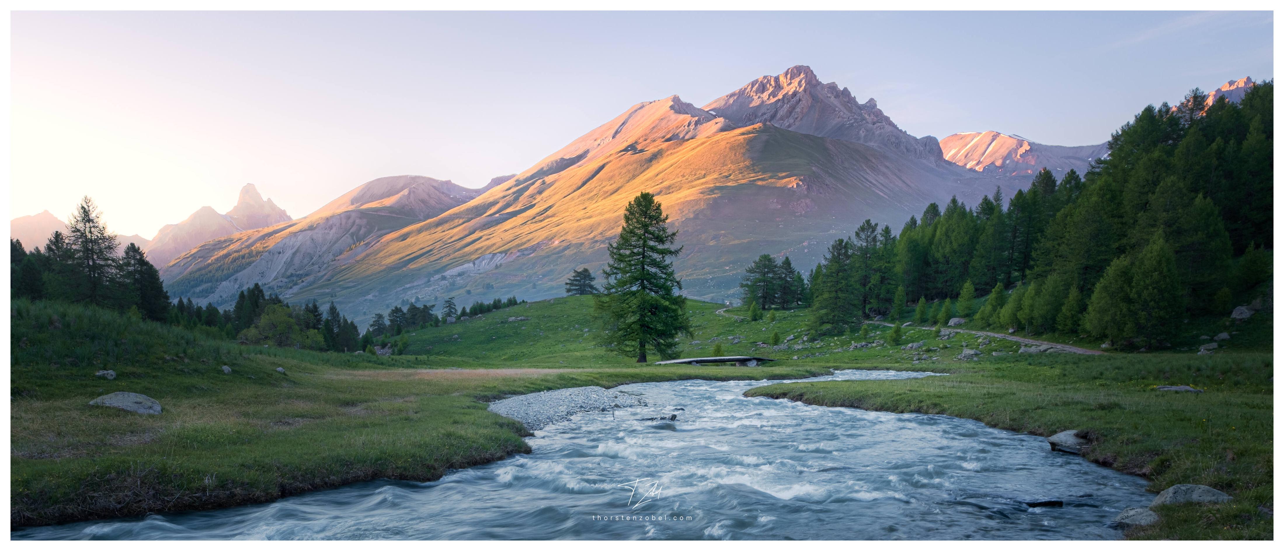 A river flows through a flat and green valley. On the horizon stretches a mountain whose meadows and stones are bathed in an orange light by the setting sun.