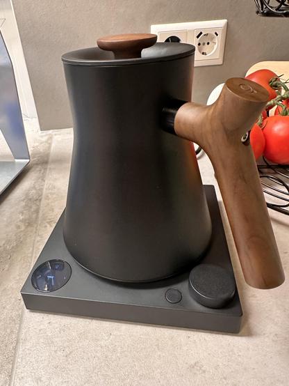 Moderne Küchenhelfer: Ein schwarzer Wasserkocher mit Holzgriff steht bereit auf seiner Basis. Das Design ist schlicht und elegant, mit einem digitalen Display und Bedienknöpfen. Im Hintergrund sieht man eine Steckdose, frische Tomaten und ein Schneidebrett – alles bereit für eine gemütliche Tasse Tee oder Kaffee.