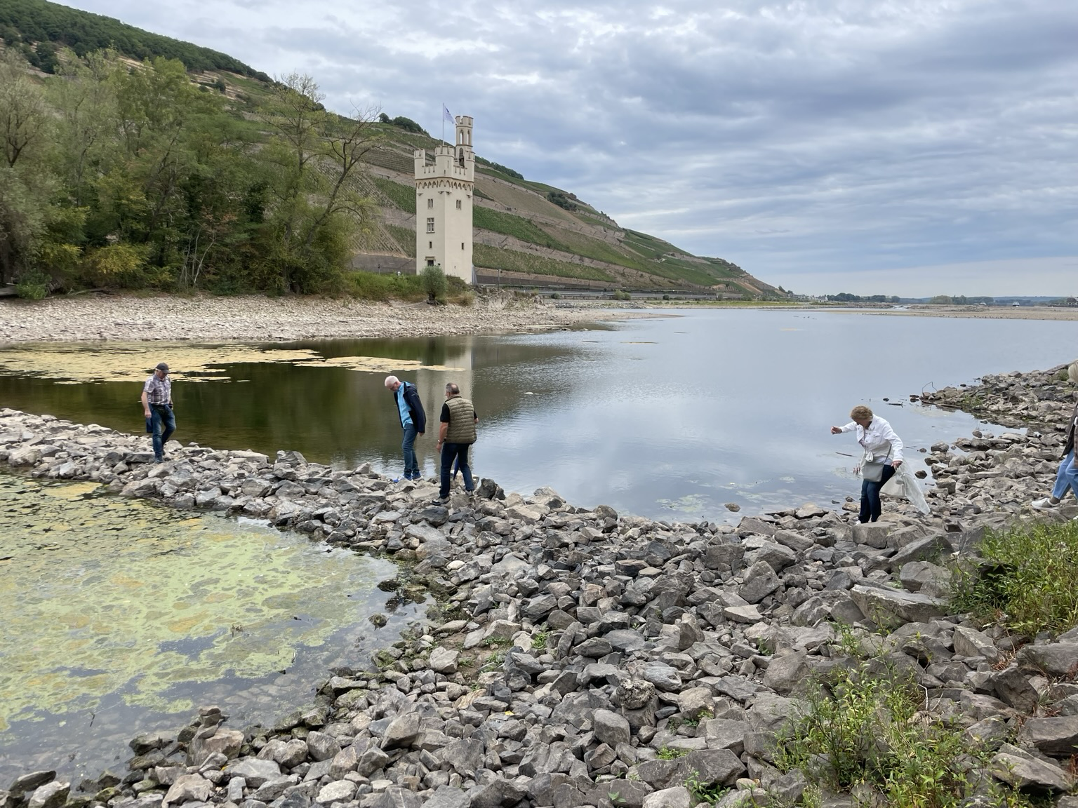 Farbiges Querformat. Flußtal mit wenig Wasser, so dass eine Insel mit einem weißen Turm fußläufig erreichbar ist. Einige Menschen gehen über große Steine.