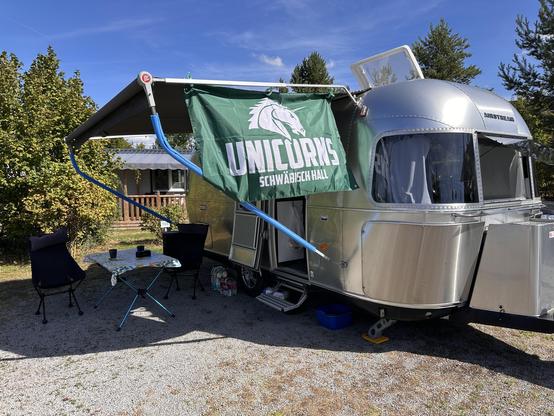 Ein silberner Airstream-Wohnwagen steht unter einem blauen Himmel, geschmückt mit der grünen Flagge der Schwäbisch Hall Unicorns. Vor dem Wohnwagen lädt ein Klapptisch mit Stühlen zum Verweilen ein. Im Hintergrund grüne Bäume und ein Holzhaus – die perfekte Mischung aus Natur und American-Football-Geist! 🏈🌲