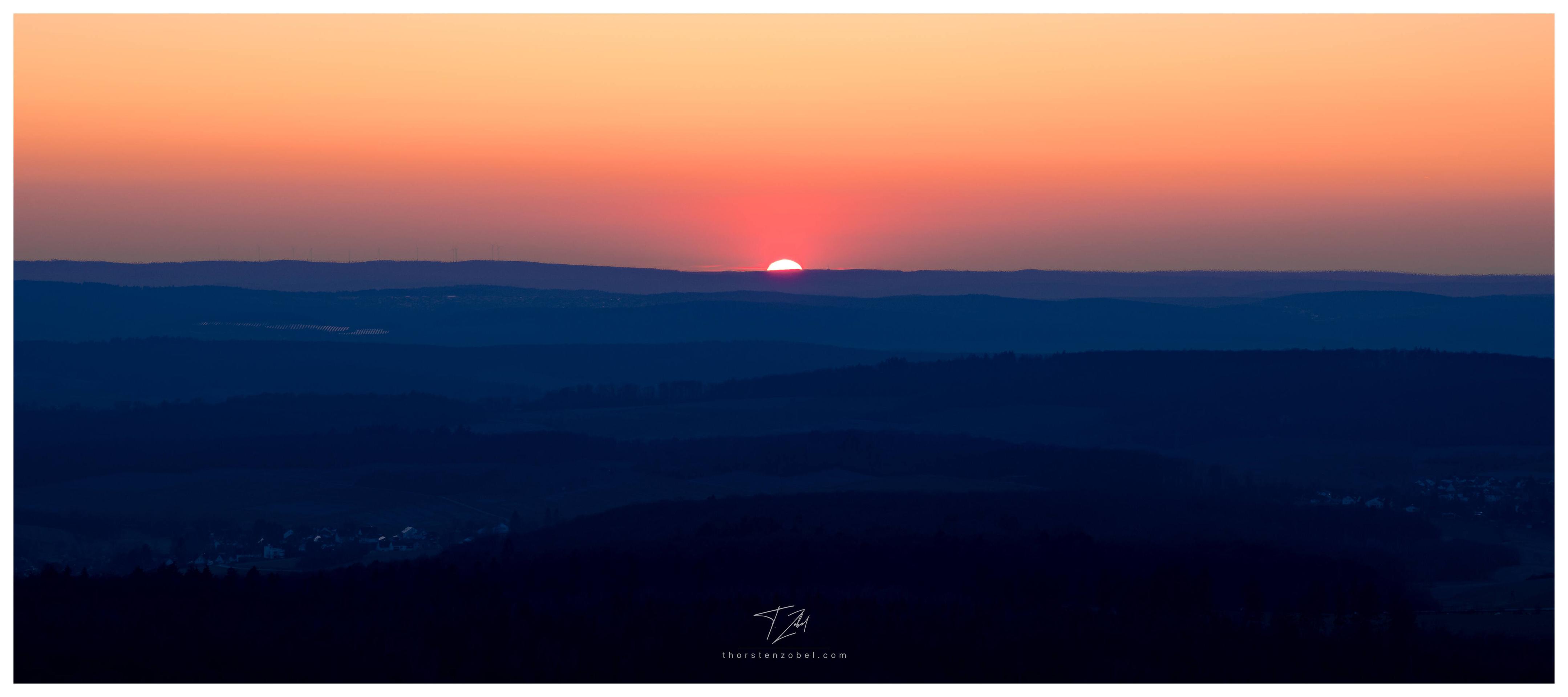 The sun sets behind a hilly landscape and is visible as half a ball of fire on the horizon. The hilly landscape has various shades of dark blue, while the sun bathes the sky in a warm orange-red light.