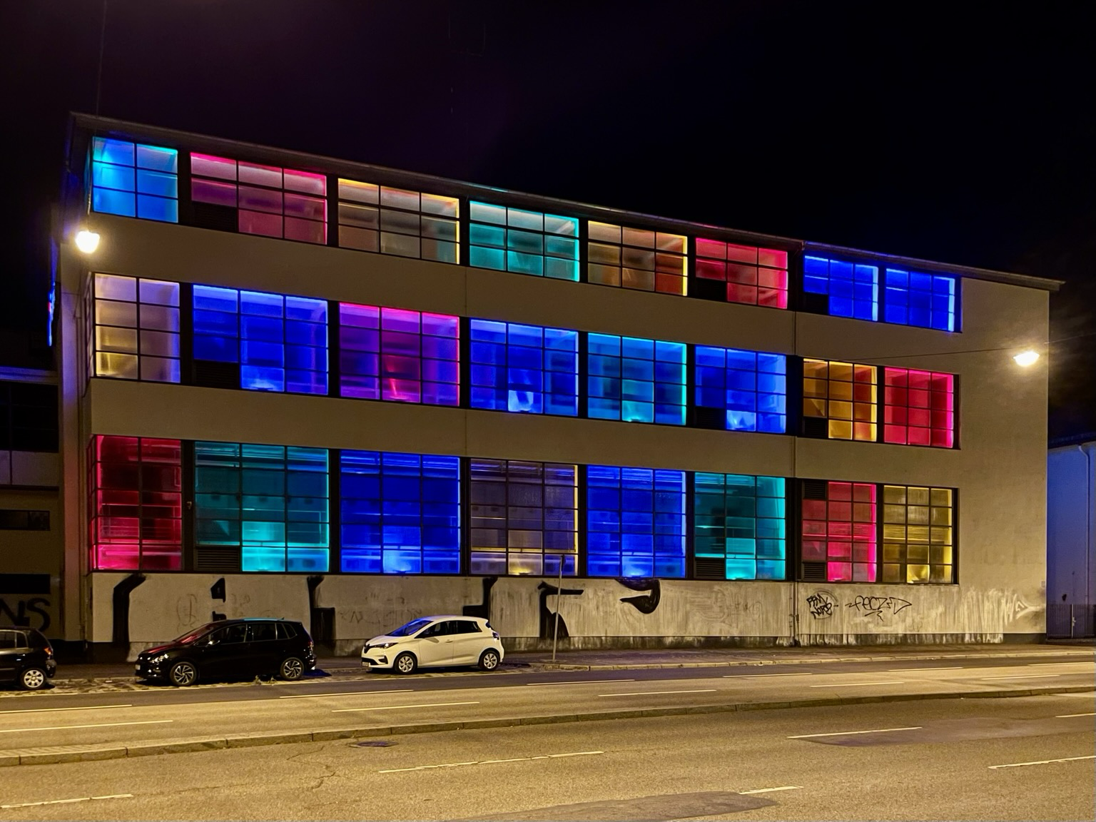 Farbiges Querformat. Dreistöckiges Gebäude an einer Straße bei Nacht. Die großen Fenster sind bunt erleuchtet.