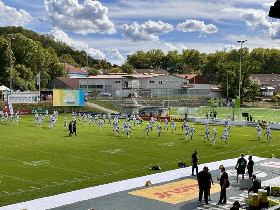 Ein American-Football-Feld auf einem grünen Kunstrasenfeld unter klarem Himmel mit einigen Wolken.
- Vordergrund: Die Spieler:innen der Schwäbisch Hall Unicorns (in weißen Trikots und Helmen) sind auf dem Feld positioniert und bereiten sich auf das Spiel vor.
- Hintergrund:
- Eine Anzeigetafel mit der Aufschrift „Indoor‘s“ und weiteren nicht lesbaren Inhalten.
- Tribünen mit wenigen Zuschauer:innen, umgeben von Werbebannern, darunter „Gengenbach“ und „Sparkasse“.
- Gebäude und Bäume sind hinter…