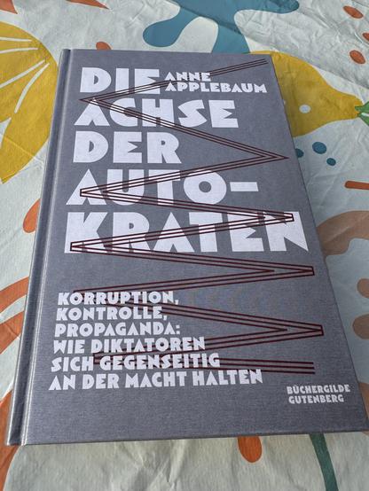 Ein Buch mit dem Titel „Die Achse der Autokraten“ von Anne Applebaum liegt auf einem bunten Hintergrund mit abstrakten, organischen Formen in Gelb, Blau, Orange und Weiß.
- Titel: „DIE ACHSE DER AUTO-KRATEN“ in großen, weißen Buchstaben auf dem Einband.
- Untertitel: „Korruption, Kontrolle, Propaganda: Wie Diktatoren sich gegenseitig an der Macht halten“ in kleinerer, weißer Schrift.
- Autorin: Anne Applebaum (Name in weißer Schrift oben auf dem Einband).
- Verlag: Buchergilde Gutenberg (Logo u…