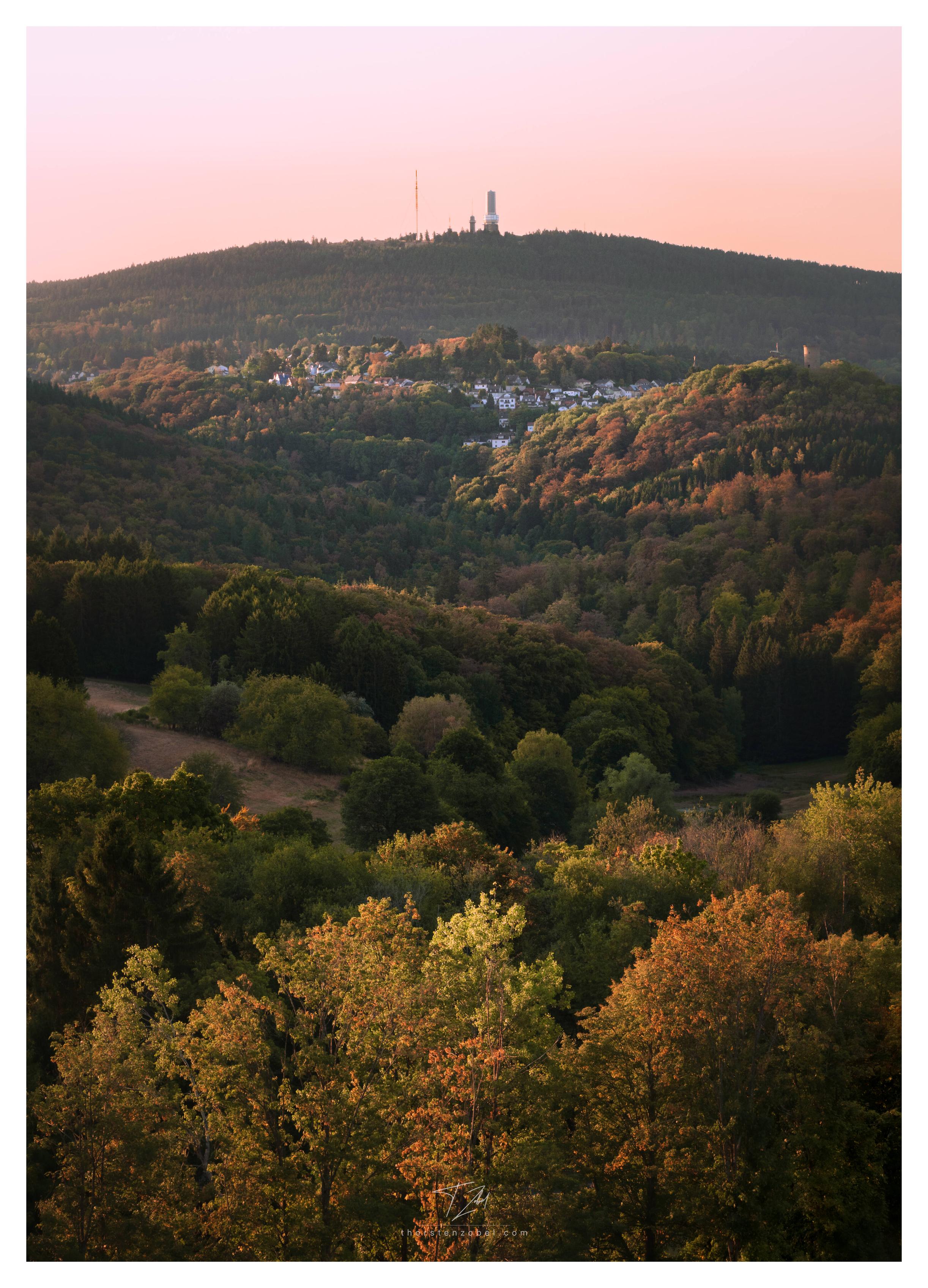 The sun illuminates the slopes of a gentle, wooded valley that stretches toward the horizon toward a larger mountain. Several towers and an antenna are visible on the mountain. A town can be seen in front of the mountain. The ruins of a castle rise on one of the hills along the valley.