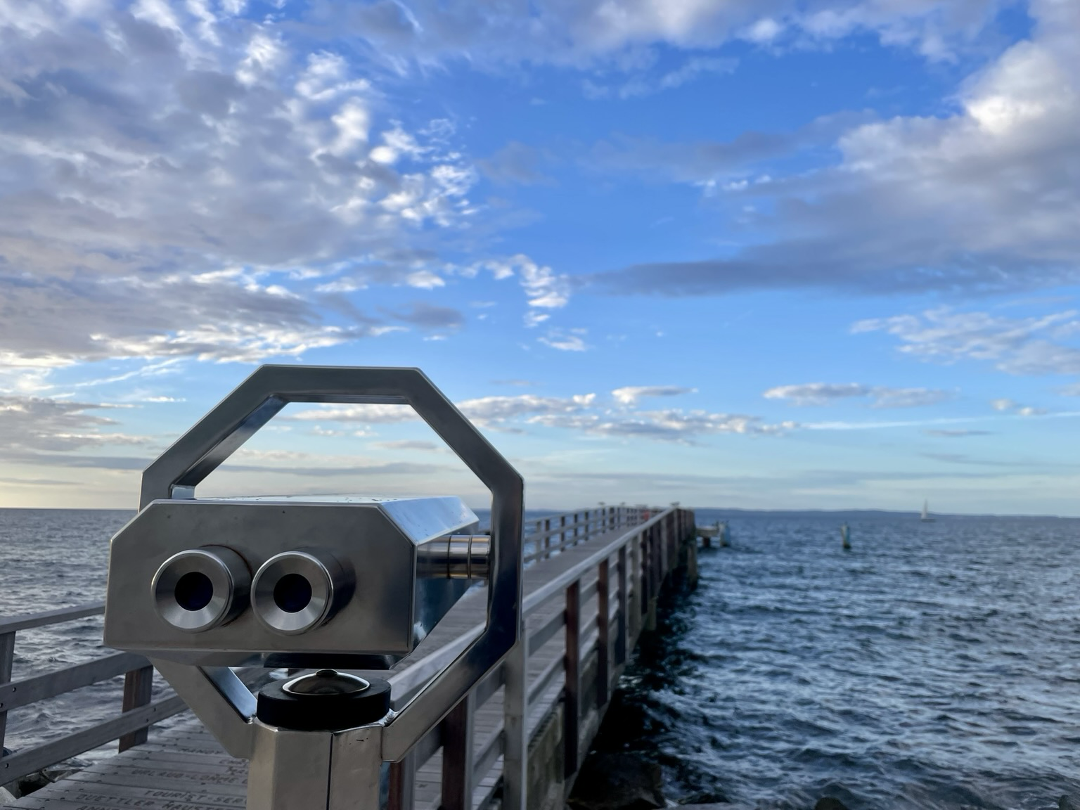Farbiges Querformat. Himmel mit Wolken, Meer, Steg und fest montiertes Fernglas, das aussieht wie ein Marsianer in einem billig gemachten Science Fiction Film oder einem KI generierten Video zur Erläuterung des Meeresleuchtens.