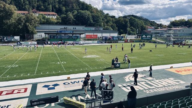 Ein American-Football-Spielfeld vor dem Spiel der Schwäbisch Hall Unicorns bei bewölktem Himmel.
- Vordergrund:
- Das grüne Spielfeld mit weißen Linien für die Yard-Markierungen.
- Spieler:innen in weißen und grünen Trikots sind auf dem Feld verteilt.
- An der Seitenlinie stehen Betreuer:innen, Schiedsrichter:innen und Teammitglieder in schwarzer Kleidung.
- Ein Tisch mit Ausrüstung und ein Wasserbehälter sind am Rand des Feldes zu sehen.
- Hintergrund:
- Tribünen mit Werbebannern, darunter „GW…