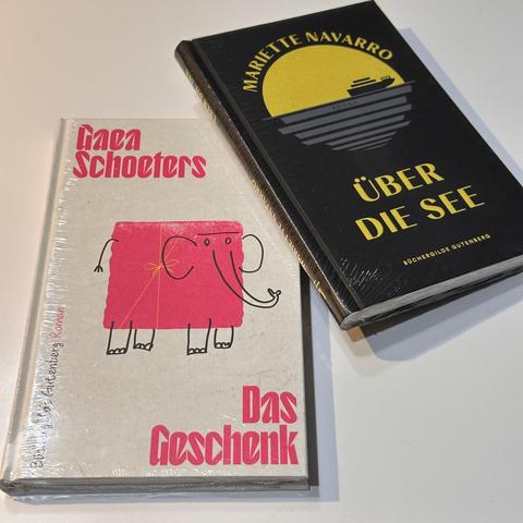 Zwei Bücher liegen nebeneinander auf einer hellen Oberfläche, beide noch in durchsichtiger Plastikfolie eingewickelt.
- Linkes Buch:
- Titel: „Das Geschenk“ von Gaea Schoeters (Autorenname in roter Schrift, Titel in Rot und Schwarz).
- Cover: Grauer Einband mit einer Illustration eines pinken Elefanten, der ein Geschenk auf dem Rücken trägt.
- Rechtes Buch:
- Titel: „Über die See“ von Mariette Navarro (Texte in gelber Schrift).
- Cover: Schwarzer Einband mit einer grafischen Darstellung eines S…