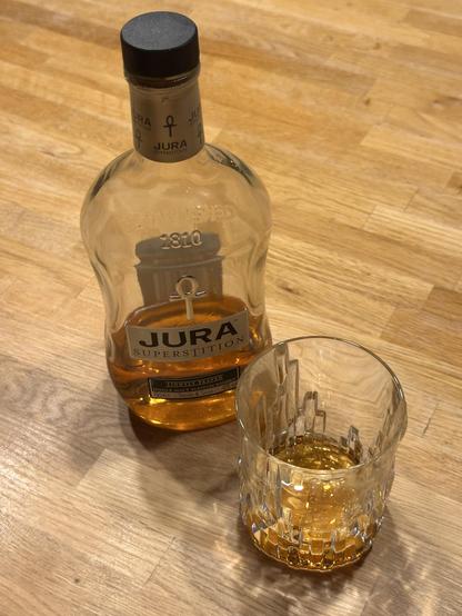 Eine Flasche Jura Superstition Whisky und ein Glas mit Whisky auf einem hellen Holztisch.
- Flasche: Klares Glas mit schwarzem Schraubverschluss, goldfarbene Flüssigkeit. Lesbare Texte: „Jura“, „Superstition“, „Established 1810“ und ein ankerähnliches Symbol.
- Glas: Klares Glas mit facettiertem Design, enthält eine kleine Menge goldfarbenen Whiskys.
- Hintergrund: Heller Holztisch.
Die Stimmung wirkt ruhig und gemütlich – ein entspannter Whiskyabend.